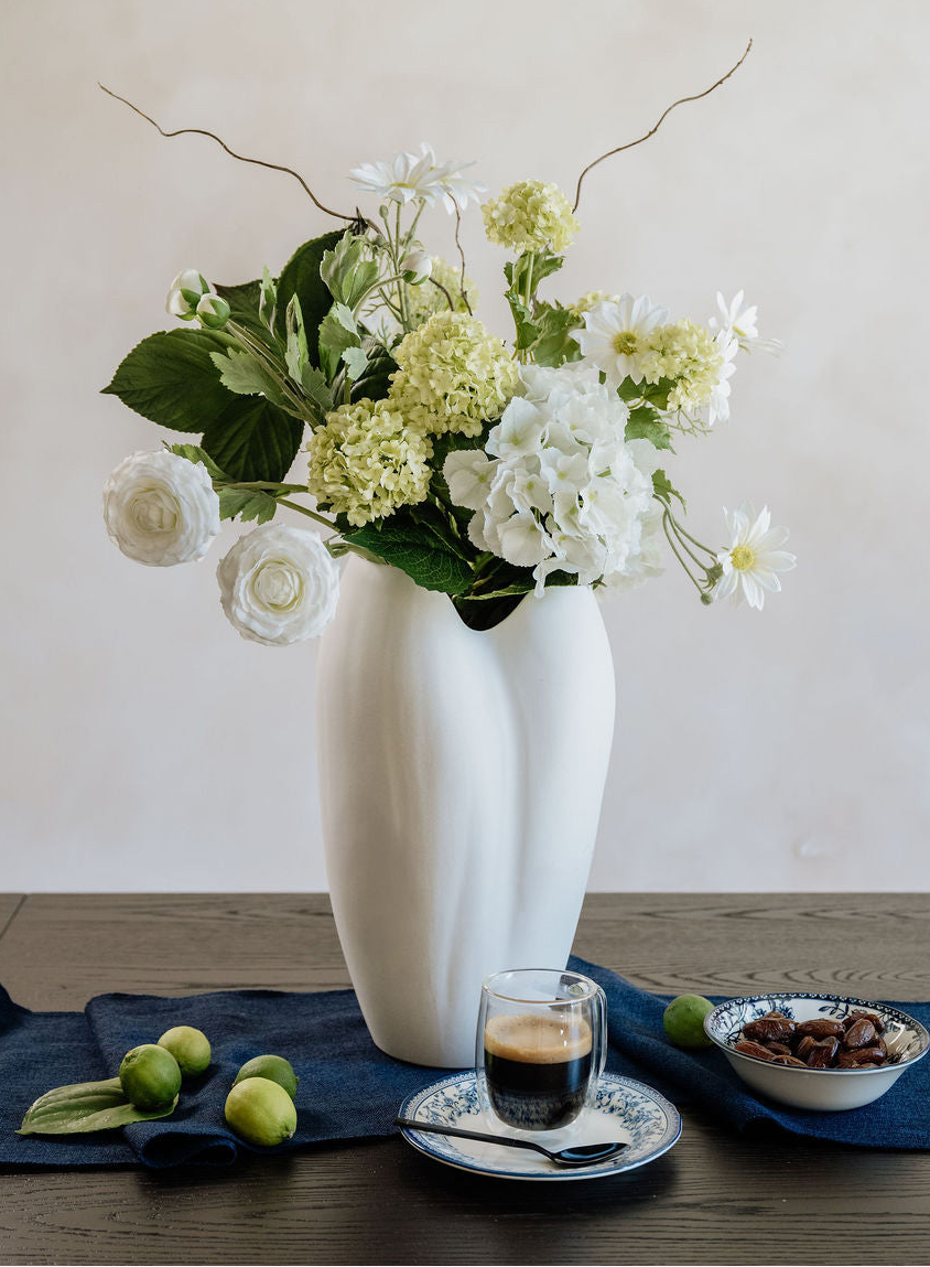 Vase with faux flowers on a table with a cup of coffee and snacks.